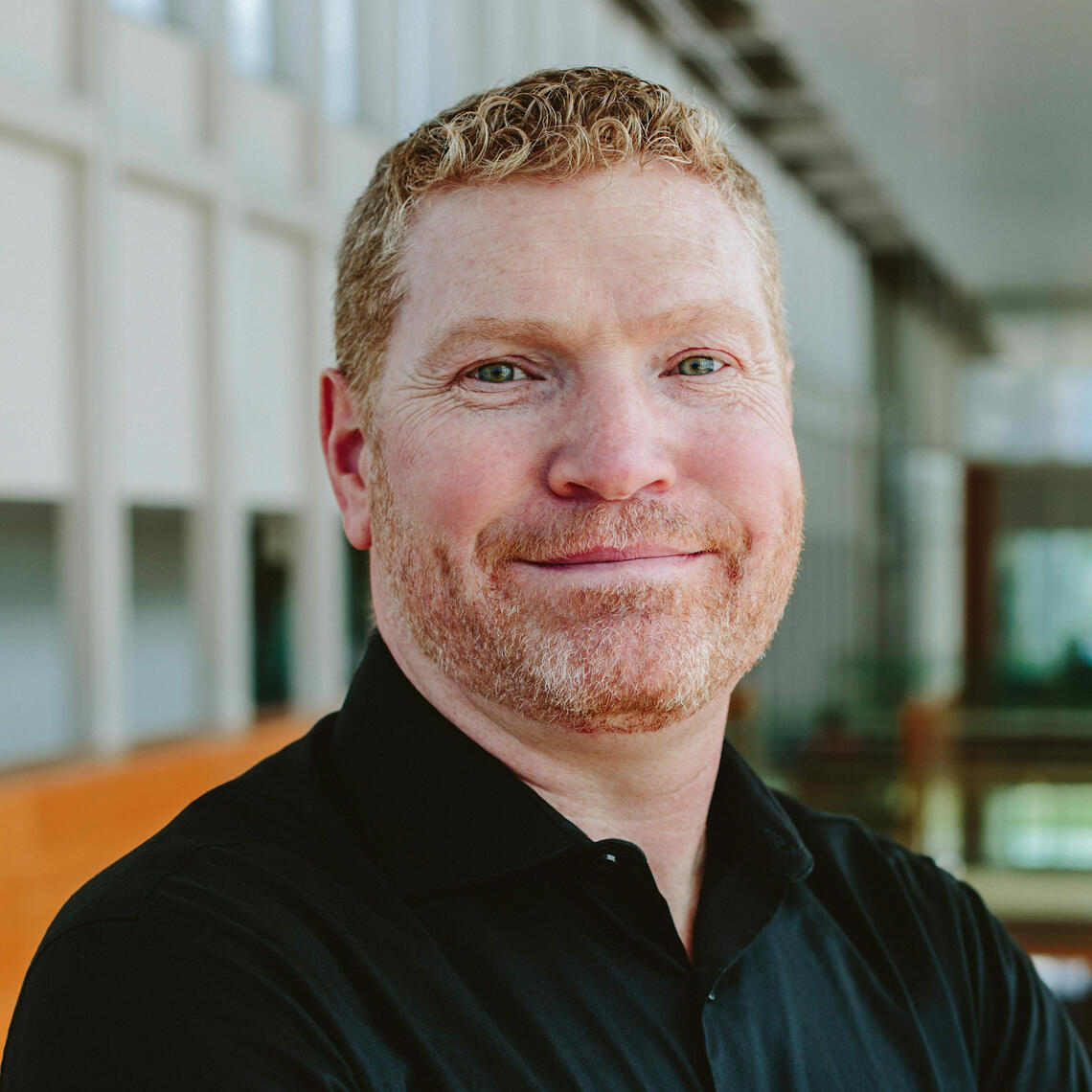 Corey Flynn, a light-skinned man with ginger hair and beard, smiles inside the Taylor Institute.